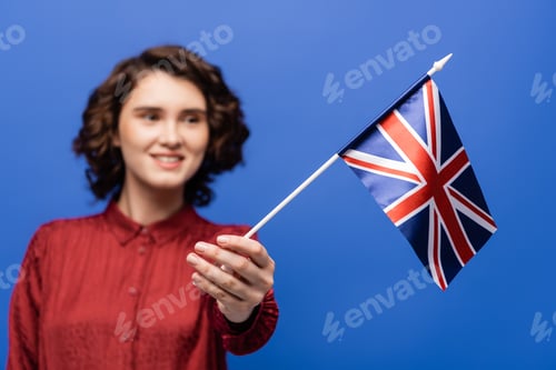 Preview: joyful student with curly hair looking at flag of United Kingdom isolated on blue