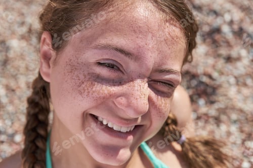Preview: Happy teenage girl with freckles on her face and pigtails enjoys the sun while sitting on the beach.