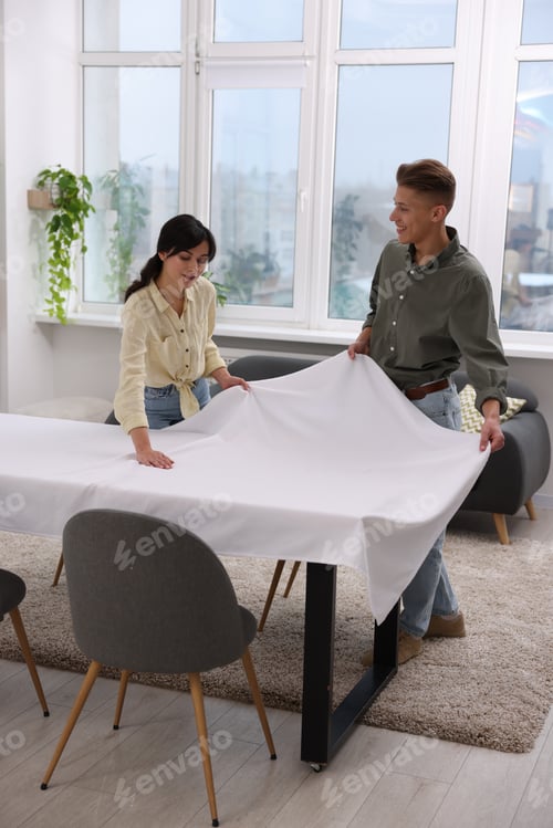 Preview: Couple putting white tablecloth on table at home