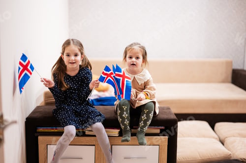 Preview: Two sisters are sitting on a couch at home with icelandic flags on hands.