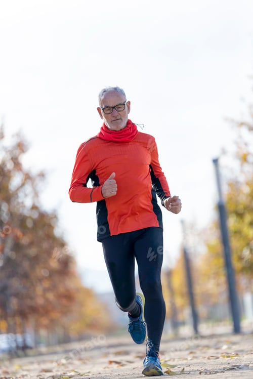 Preview: Front view of a senior man in sport clothes jogging in a city park in a sunny day