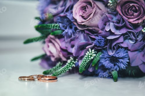 Preview: Romantic Wedding Rings on Table Beside Elegant Floral Bouquet of Purple Roses and Blossoms