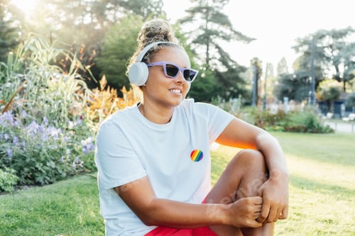 Preview: Smiling mixed race young woman with a rainbow badge LGBTQ listening music with wireless headphones