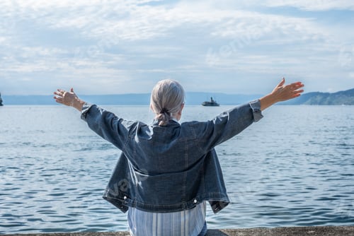 Preview: Rear view of senior white haired woman sitting along lake with open arms admiring landscape