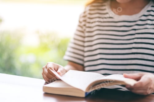Preview: Closeup image of a woman holding and reading a vintage novel book on wooden table