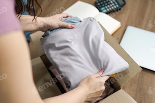Preview: Woman Packing Clothes into a Cardboard Box for Moving or Donation on a Wooden Table with Calculator
