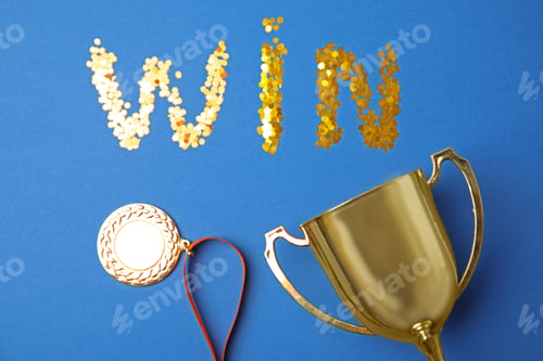 Preview: Gold trophy cup, medal and word WIN made with confetti on blue background, flat lay
