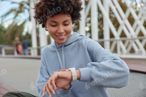 Preview: Attractive positive young Afro American woman checks time on wrist watch glad not to be late on meet