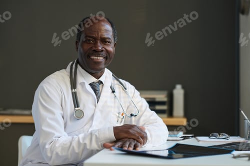 Preview: Successful mature doctor in lab coat sitting by workplace in clinics