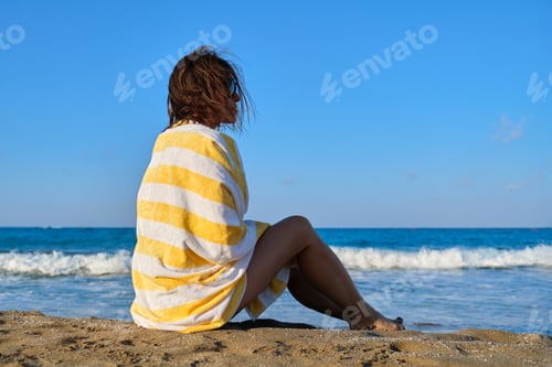 Preview: Mature woman sitting on seashore, resting beautiful female with beach towel on her shoulders