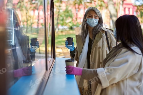 Preview: Elegant women in medical masks drinking coffee and chatting outdoors
