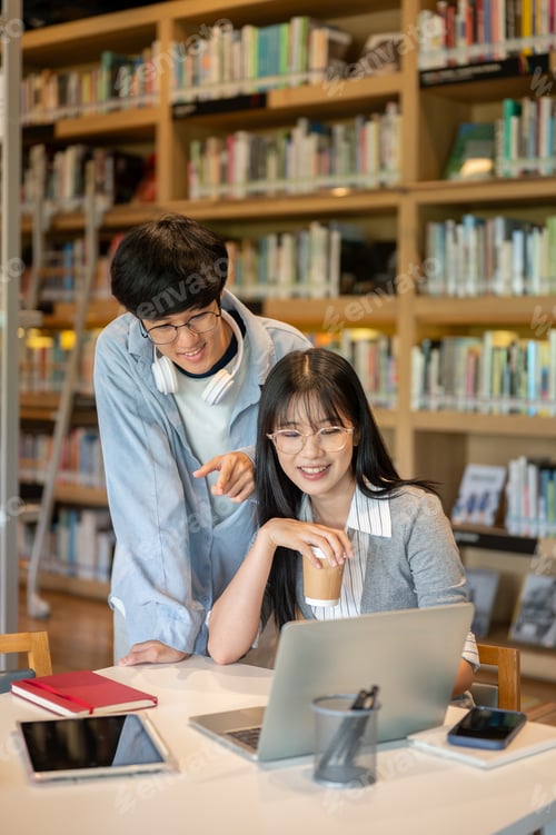 Preview: Glasses asian man student standing behind woman friend pointing looking at laptop on library's table