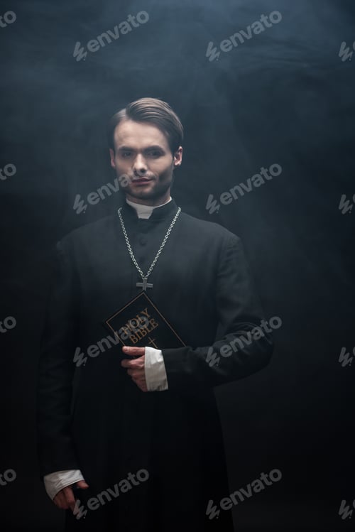 Preview: confident catholic priest holding holy bible and looking at camera on black background with smoke