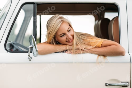 Preview: Smiling young beautiful blond woman leaning out from car window looking away