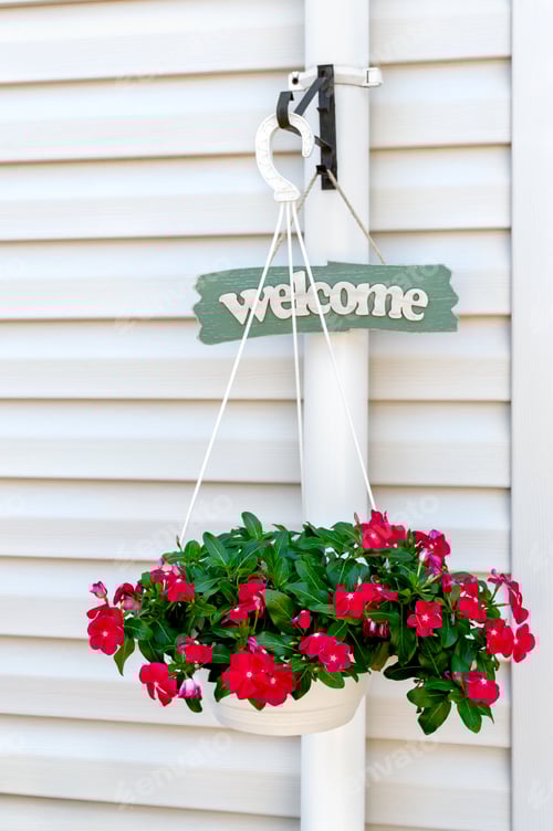 Preview: Board with "Welcome" sign and beautiful flowers in a pot near the white wall of the house.
