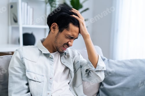 Preview: Asian young upset depressed man sitting alone in living room at home.