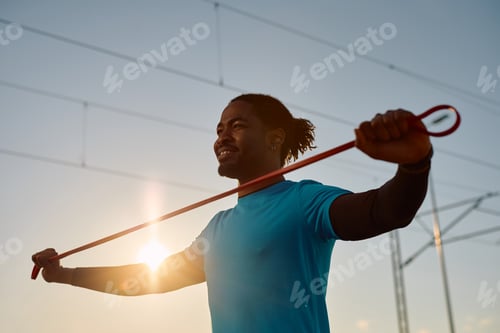 Preview: Black athletic man using resistance band while working out at sunset.