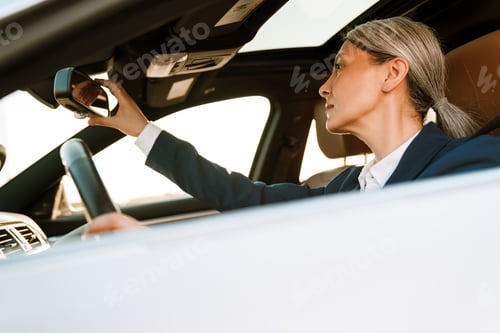 Preview: Mature asian businesswoman looking in rear-view mirror while driving car
