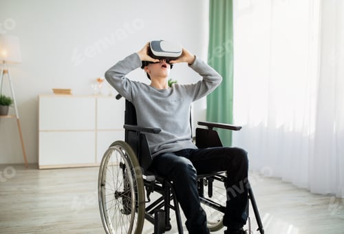 Preview: Happy handicapped teen boy in wheelchair using VR headset to explore augmented reality at home