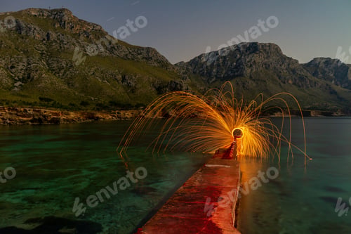 Preview: Person with burning circle on stones between water near hills at night
