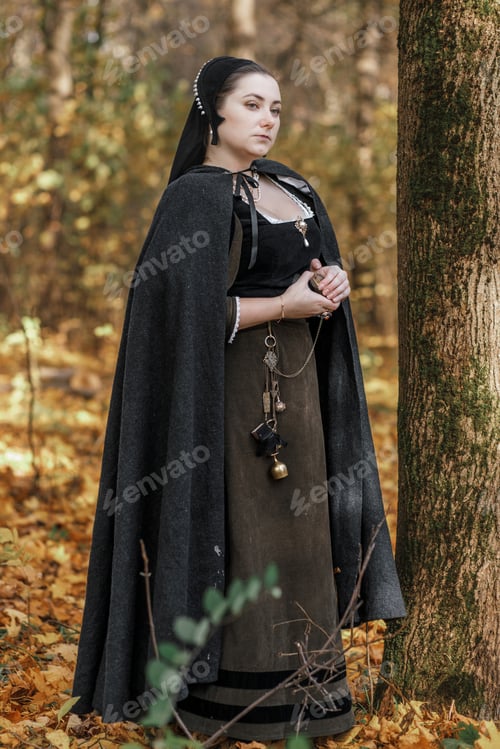 Preview: Young woman in medieval dress and cloak standing by tree in forest. Middle Ages