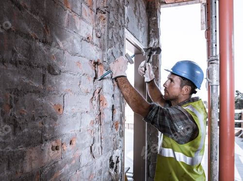 Preview: A worker opens a hole in a brick wall using a chisel and hammer in a building under construction.