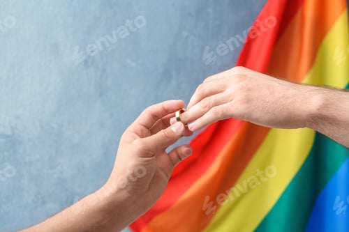 Preview: Young man putting wedding ring on his boyfriend's finger near rainbow flag. Gay marriage