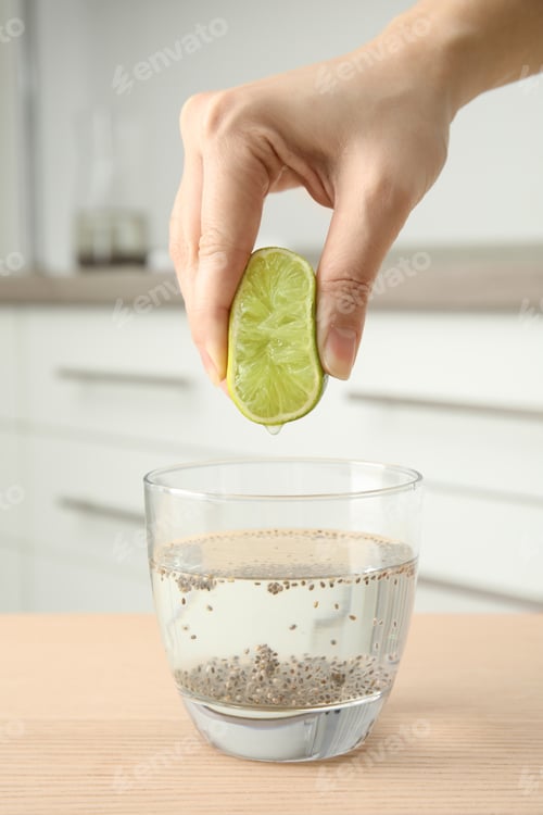 Preview: Young woman squeezing lime into glass of water with chia seeds on table, closeup