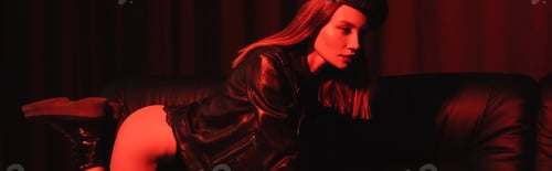 Preview: sexy woman in leather jacket and boots posing on couch in red light on black background, banner