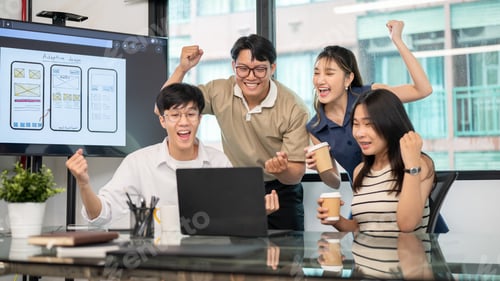 Preview: Asian man and coworkers excitedly raising fist while looking at laptop on glass top table in office