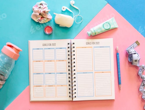 Preview: Overhead view of a planner open to goals pages, earbuds, hair scrunch, lip gloss, water bottle, pen