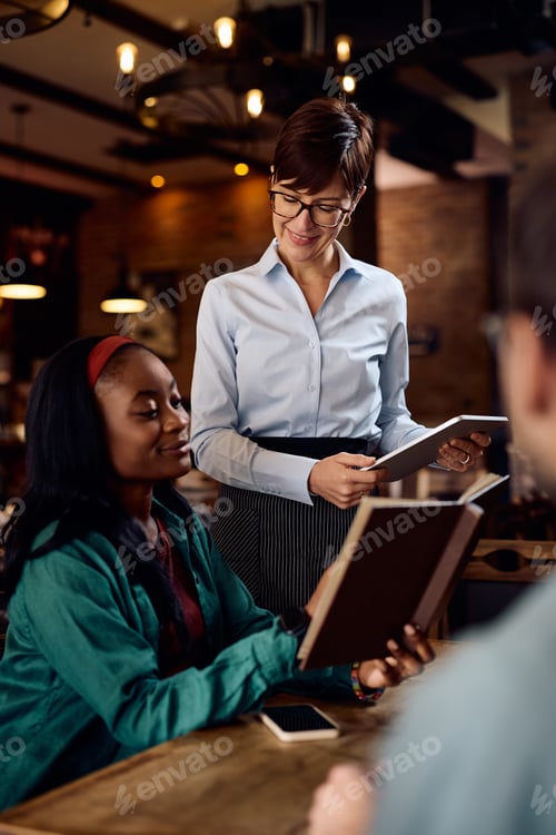 Preview: Happy waitress using digital tablet while taking order from black woman in a bar.