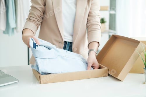 Preview: Asian female clothes shop owner folding a t-shirt and packing in a cardboard parcel box.