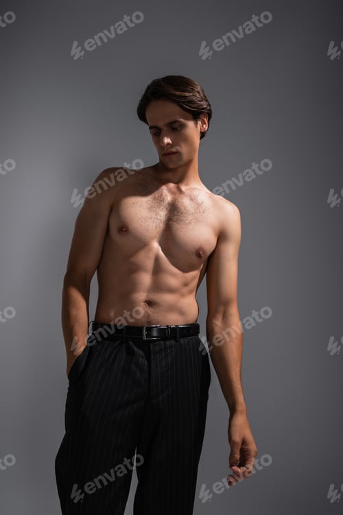Preview: good looking man with muscular body posing with hand in pocket of black trousers on grey