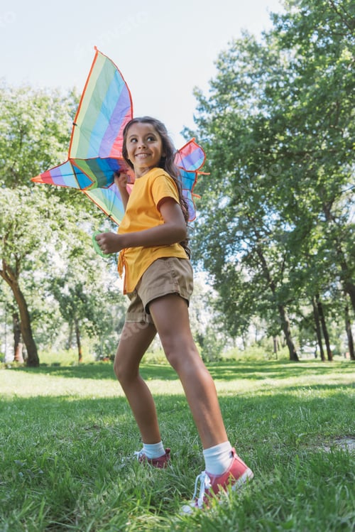 Preview: low angle view of cute happy child holding colorful kite and looking away in park