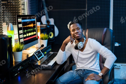 Preview: Young successful African man in casualwear sitting by workplace in studio