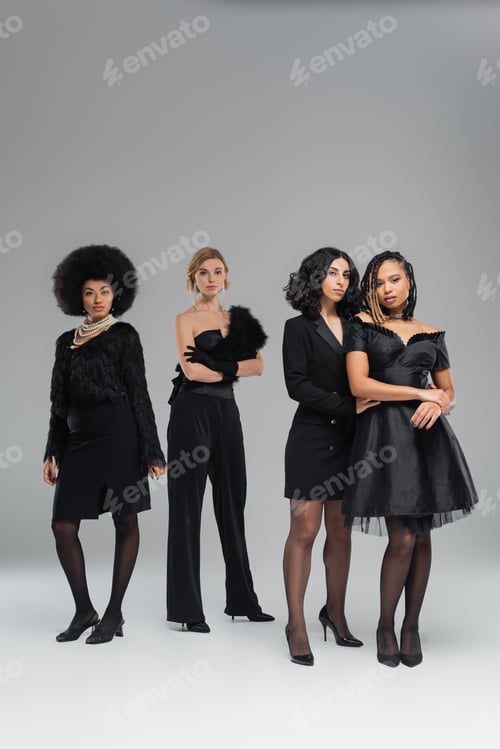 Preview: full length of glamorous fashionable multiracial girlfriends in black attire standing on grey