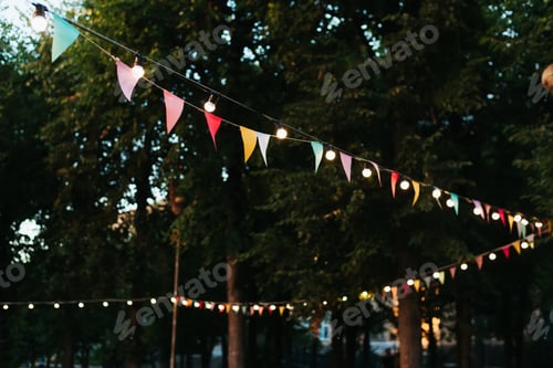 Preview: Multicolored decorative festival flags and light bulbs garland on street on summer evening.