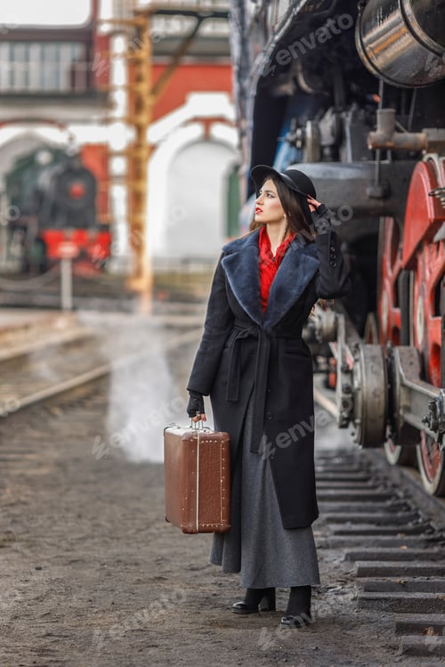 Preview: A girl in a black coat and hat with a suitcase near an old steam locomotive.Vintage portrait