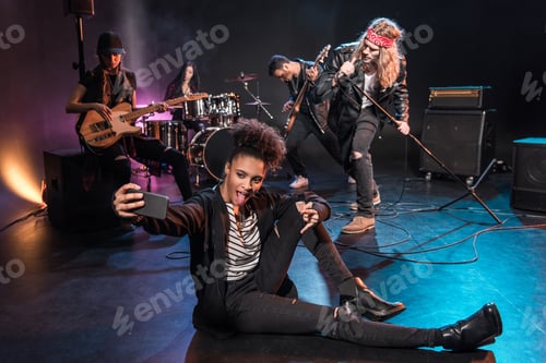 Preview: Young woman taking selfie with rock and roll band performing concert on stage