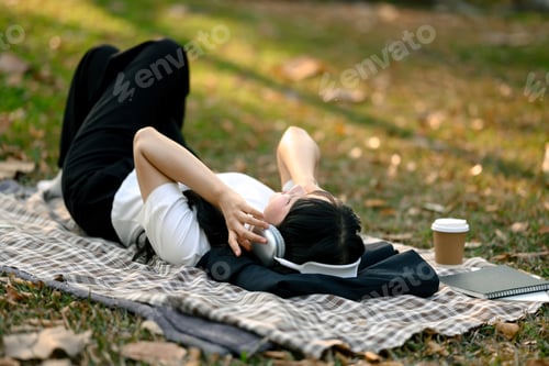 Preview: Above view of happy female office worker listening to music in headphones, lying on blanket.