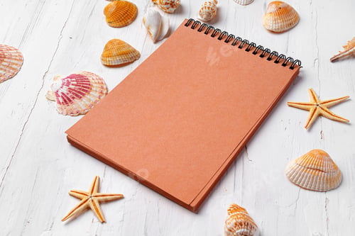 Preview: Brown open notepad surrounded by sea shells on white wooden table closeup