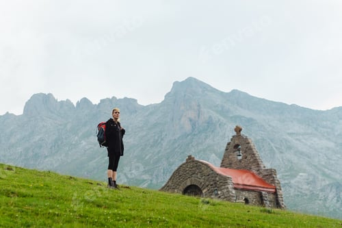 Preview: backpacking hiker hiking, ascending a green slope of the mountain valley and near a church.