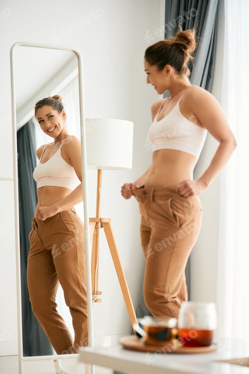 Preview: Young woman looking herself in a mirror and feeling satisfied after loosing weight.