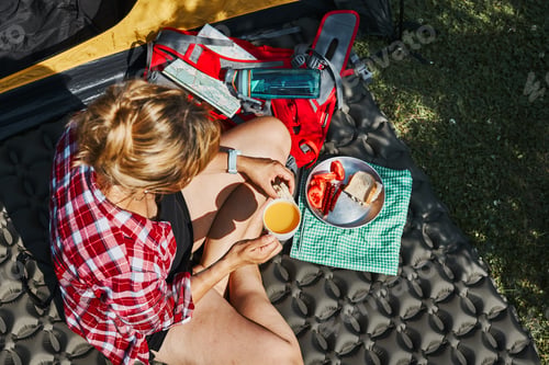 Preview: Woman having breakfast sitting by tent at camping. Female actively spending summer vacations