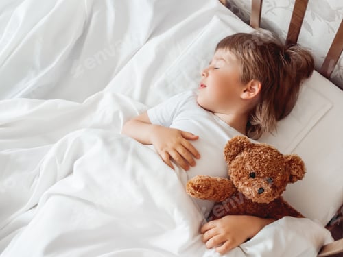 Preview: Toddler sleeps in bed with cute teddy bear. Little boy under white blanket with fluffy toy.