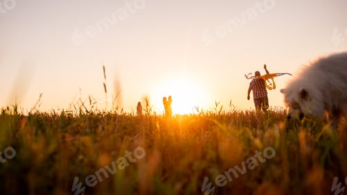 Preview: Defocused panoramic diversity family with Samoyed running at sunset with kite in sky.
