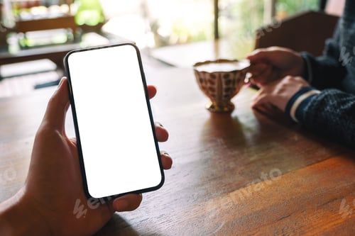 Preview: a man's hand holding black mobile phone with blank white screen with woman drinking coffee in cafe
