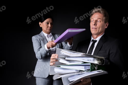 Preview: frustrated mature businessman holding pile of folders while female colleague putting folder on the