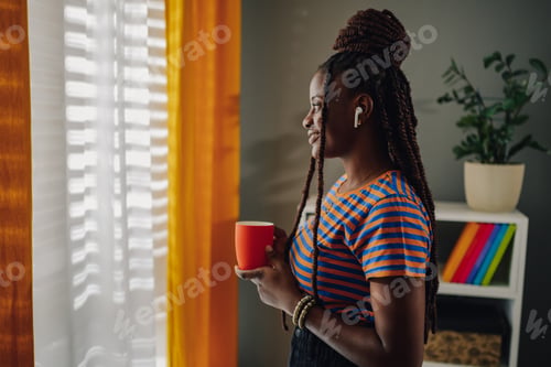 Preview: Young woman enjoying a cup of coffee while listening to music by the window